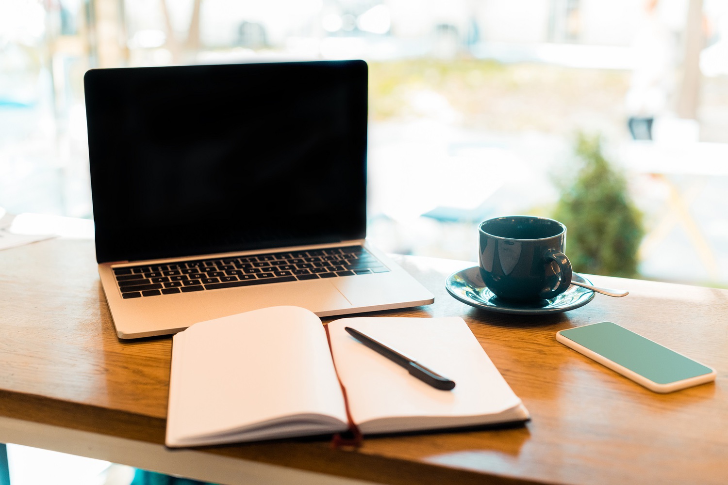 Silver laptop, open notebook with pen, dark blue coffee cup on saucer, and white smartphone on a wooden table